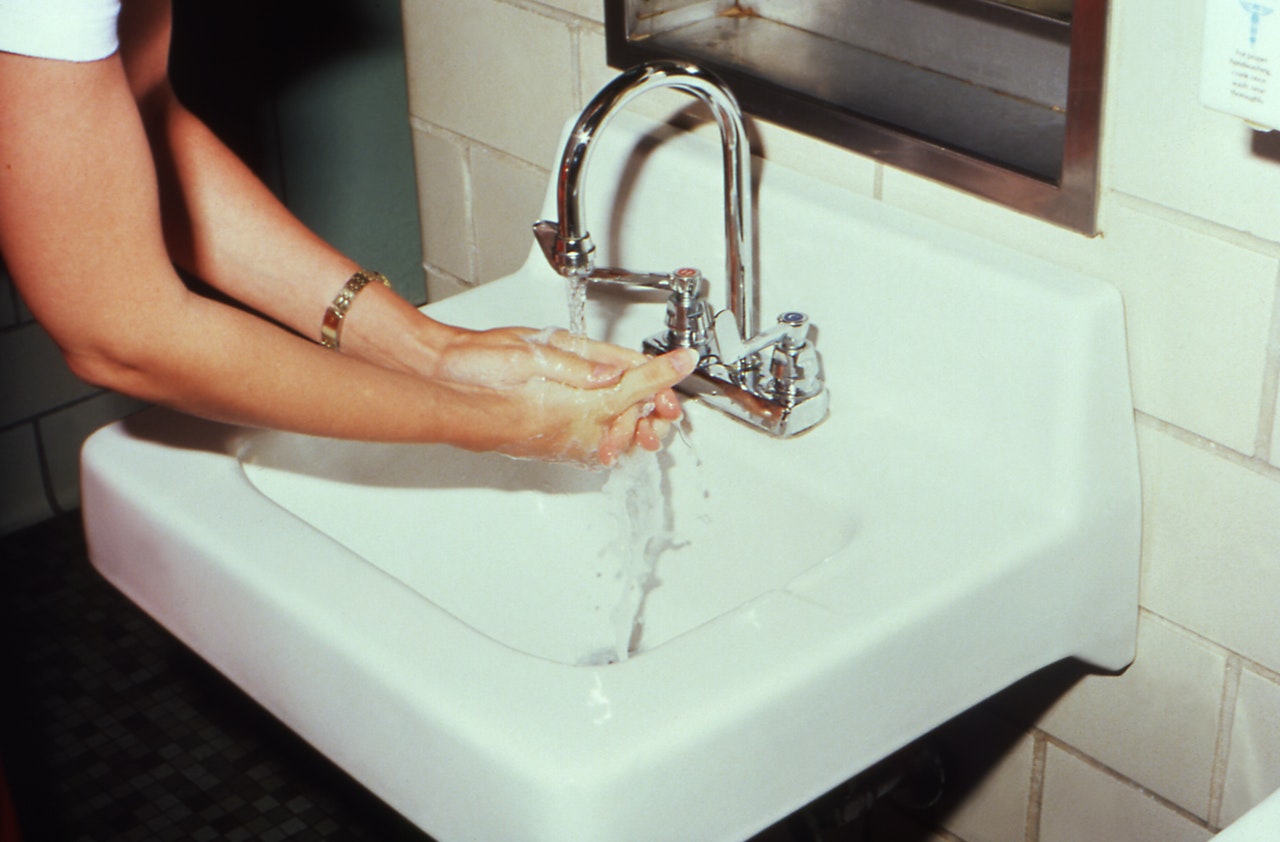 A woman in a white t-shirt washes her hands at a small basin