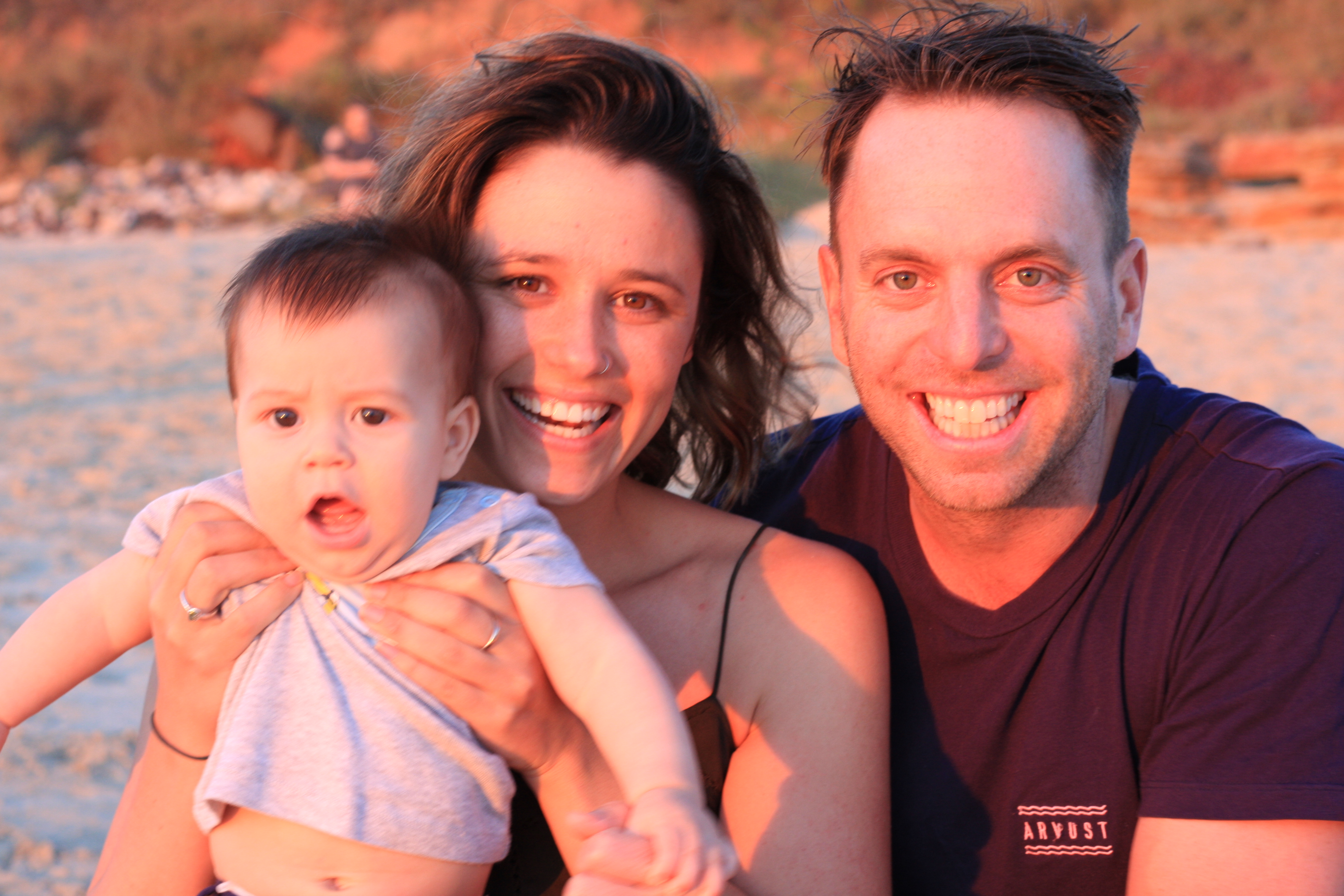 A white man and woman with dark hair sit smiling on a beach, holding a young child